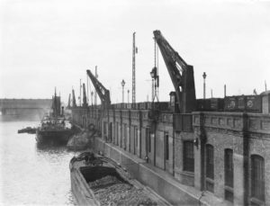 Cranes atop the brick walls of the dock, with two ships in the channel below.